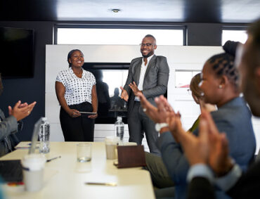 Group of mixed race colleagues in modern meeting room with laptop computer encouraging two attractive African American business professionals leading a collaborative discussion